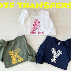 May include: Three crewneck sweatshirts featuring distressed varsity-style letters. A white sweatshirt with a pink letter A, an olive green sweatshirt with a yellow letter K, and a navy blue sweatshirt with a white letter Y. Text reads DTF TRANSFERS.