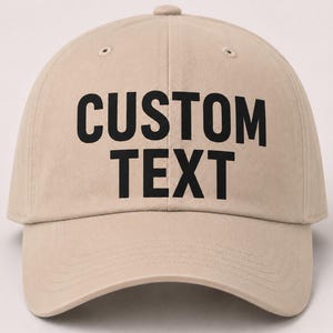May include: A beige baseball cap featuring the words CUSTOM TEXT printed in bold black capital letters across the front panels. The cap has a curved brim, a button on the top center, and stitched eyelets for ventilation on each side panel.