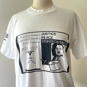 Puede incluir: Camiseta blanca con un estampado gráfico en blanco y negro de un hombre tocando la batería. El gráfico incluye el texto "Justicia Paz Rectitud" y "Drum Major Award".