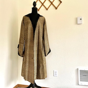 Könnte beinhalten: Ein langer, gold-schwarz gemusterter Mantel mit schwarzem Kragen und Ärmelbündchen. Der Mantel befindet sich auf einem Mannequin und hat eine lockere Passform.
