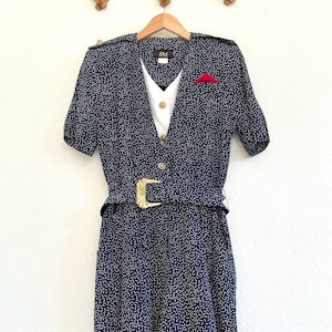 Puede incluir: Un vestido azul marino con lunares blancos y una camisa blanca con cuello debajo. El vestido tiene un cinturón con hebilla dorada y un pin rojo en el pecho.