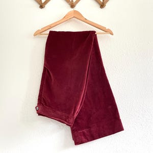Puede incluir: Un par de pantalones de pierna ancha de color burdeos oscuro, colgados de una percha de madera contra una pared blanca.