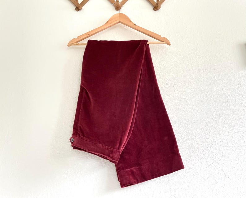 Puede incluir: Un par de pantalones de pierna ancha de color burdeos oscuro, colgados de una percha de madera contra una pared blanca.