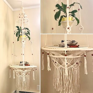 Può includere: Un ripiano sospeso in macramè bianco con un piano in vetro e perline di legno. Il ripiano è appeso al soffitto e ha una pianta appesa ad esso.