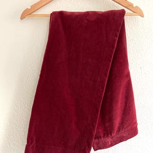 Puede incluir: Un pantal&oacute;n de terciopelo burdeos colgado de una percha de madera.