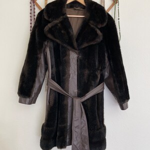 May include: A brown faux fur coat with a leather belt and a leather panel down the front. The coat has a collar and long sleeves.