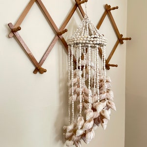 May include: A white seashell wind chime hanging from a wooden wall rack. The wind chime is made of multiple strands of white beads and seashells.