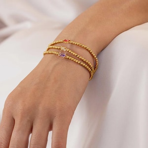 May include: Gold beaded bracelets with rectangular gemstones in red, yellow, and purple. The bracelets are stacked on a wrist. The person is wearing a white dress. The bracelets are made of small gold beads.