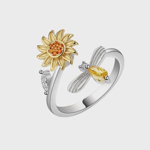 May include: A silver-toned open ring featuring a gold-toned sunflower with an orange center on one end and a silver-toned bee with a yellow teardrop-shaped stone body and a small clear stone on its head on the other end of the band.