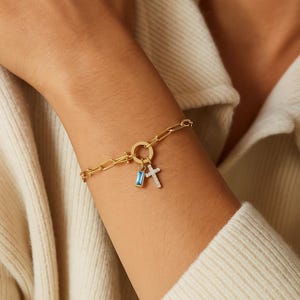 Könnte beinhalten: Goldfarbenes Kettenarmband mit einem rechteckigen blauen Anhänger und einem Kreuzanhänger. Das Armband besteht aus Kettengliedern und hat einen runden Verschluss. Die Anhänger sind an einem runden Verbinder befestigt.