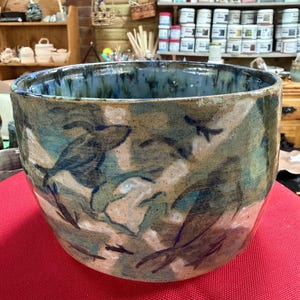 May include: A large, handmade ceramic bowl with a wide opening. The exterior features a camouflage-like pattern in shades of blue, green, and tan. The rim has a glossy, dark blue glaze. The bowl sits on a red surface.
