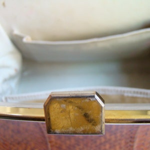 Puede incluir: Primer plano del interior de un bolso vintage con un cierre de metal dorado. El bolso est&aacute; forrado con una tela blanca y tiene un exterior de madera marr&oacute;n.