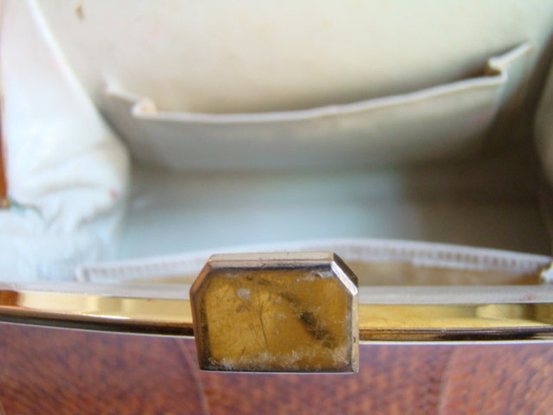 Puede incluir: Primer plano del interior de un bolso vintage con un cierre de metal dorado. El bolso est&aacute; forrado con una tela blanca y tiene un exterior de madera marr&oacute;n.