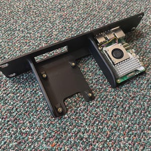 Könnte beinhalten: Schwarze elektronische Baugruppe mit Lüfter und Kühlkörper, montiert auf einer schwarzen Halterung. Die Baugruppe umfasst eine Leiterplatte und verschiedene Anschlüsse. Die Halterung ist mit Schrauben befestigt.