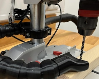 May include: A drill press with a black flexible dust collection hose and a drill bit positioned over a piece of wood. The drill press is silver and green, with a red button and a wrench attached.