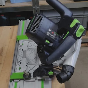 May include: A black and green power saw with a silver base. The saw has a handle and a blade guard. The base has green accents and a measuring guide. The saw is designed for cutting wood and other materials.