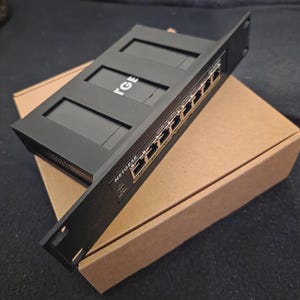 Op de afbeelding: Een zwarte Netgear netwerkswitch met meerdere poorten en de merknaam zichtbaar. De switch ligt op een bruine kartonnen doos. De bovenkant van de switch heeft de letters "TGE" erop.