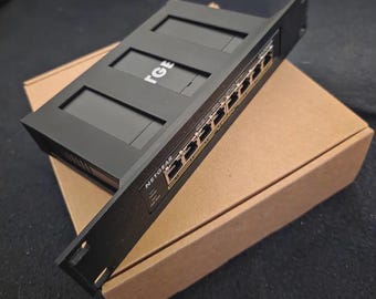 Op de afbeelding: Een zwarte Netgear netwerkswitch met meerdere poorten en de merknaam zichtbaar. De switch ligt op een bruine kartonnen doos. De bovenkant van de switch heeft de letters "TGE" erop.