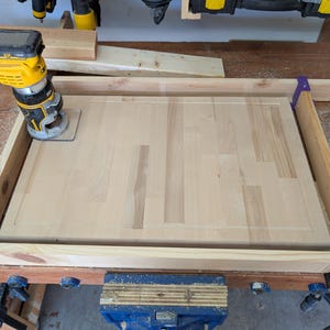 May include: A woodworking project in progress. A yellow and black router is in use on a light-coloured wooden surface. The wood is clamped within a wooden frame, with tools and sawdust visible.