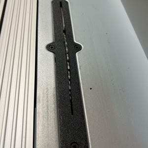 Puede incluir: Primer plano de una hoja de sierra de mesa y sus componentes. La hoja de sierra es visible dentro de un inserto negro y alargado, colocado sobre una superficie metálica plateada. El inserto está asegurado con tornillos y tiene una ranura para la hoja.