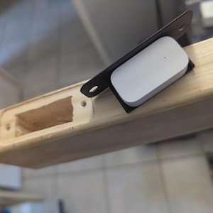 Può includere: Un dispositivo rettangolare bianco con un piccolo punto nero è posizionato su una staffa nera, posto su una tavola di legno chiaro. La tavola presenta un'incavatura rettangolare. La scena suggerisce un progetto fai-da-te o un'installazione, possibilmente legato all'elettronica o al miglioramento della casa.