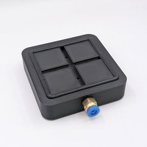 May include: A black, square-shaped object with four recessed squares on the top surface. It has a brass fitting with a blue plastic ring on one side. The object is likely a component or part of a larger system.