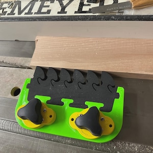 Op de afbeelding: Een groene en zwarte duwblok voor de tafelzaag met twee gele knoppen en een zwart kamachtig ontwerp. Een stuk hout is naast het duwblok op de tafelzaag geplaatst.