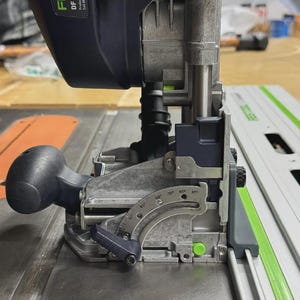 Puede incluir: Primer plano de una fresadora Festool DF 700 EQ. La herramienta es negra y gris con detalles verdes. Tiene un mango, un protector de cuchilla y una escala de medición. La herramienta está sobre una superficie metálica.