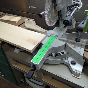 May include: A silver and black miter saw with a light wood board in place. The saw has a green guide. A small piece of wood is on the left. The saw is on a workbench.