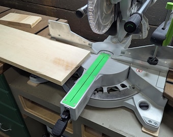 May include: A miter saw with a light wood board in place. The saw is silver and black with a green guide. A small piece of wood is on the left. The saw is on a workbench.