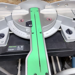 Peut inclure: Une scie à onglet argentée et noire avec un rail de guidage vert vif. La scie comporte une lame circulaire, une échelle de degrés et la marque "FESTOOL". La scie est conçue pour la coupe de précision du bois et d'autres matériaux.