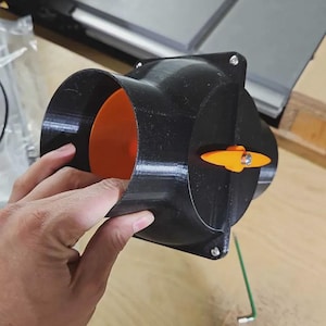 Può includere: Raccordo per la raccolta della polvere nero e arancione stampato in 3D. Il raccordo ha un ingresso cilindrico e un'uscita rettangolare con interno arancione. Sul lato è visibile una valvola a farfalla arancione. Progettato per la lavorazione del legno.