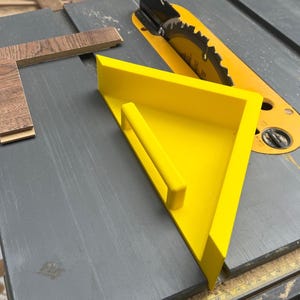 Könnte beinhalten: Ein leuchtend gelber Schiebestock mit Griff und ein hölzernes Eckstück liegen auf einer grauen Tischkreissäge. Das Sägeblatt ist sichtbar, und das Holzstück hat eine braune Maserung.