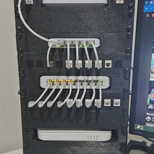 Puede incluir: Un rack de red negro lleno de conmutadores y enrutadores de red. Cables Ethernet blancos conectan los dispositivos. El rack está montado con tornillos y tiene una base de malla. Un monitor de ordenador es visible en el fondo.