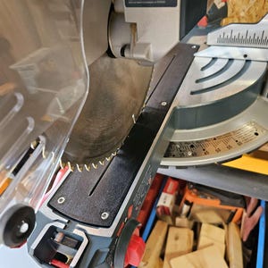 May include: A silver and black miter saw with a circular blade and a red knob. The saw has a black guide and a clear safety shield. A blue bin filled with wood scraps is in the background.