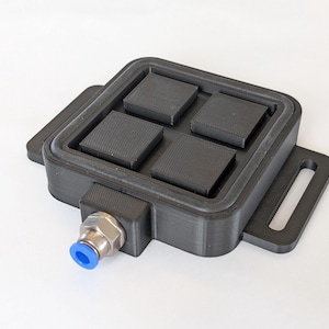 Può includere: Un dispositivo nero e rettangolare con quattro cuscinetti quadrati strutturati all'interno. Ha un telaio rettangolare con un coperchio incernierato e un raccordo con un cappuccio blu. Il dispositivo ha un'estensione a forma di maniglia sul lato.