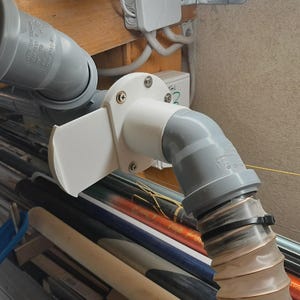 Peut inclure: Un réseau de tuyaux et raccords en plastique gris, comprenant une pièce coudée blanche avec un volet rectangulaire, relié à un tuyau flexible. Les tuyaux sont fixés avec des colliers de serrage noirs et montés sur une structure en bois.