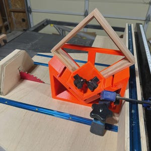 Op de afbeelding: Een oranje houtbewerkingsmal met een houten frame dat op zijn plaats is geklemd. Een rood zaagblad is zichtbaar, samen met een blauwe rail en een zwarte klem. De opstelling staat op een houten tafel.