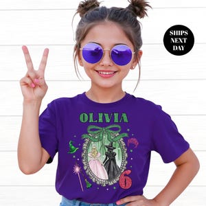 A young girl wearing a purple t-shirt with a graphic design and the text "Olivia 6" is smiling and making a peace sign.