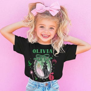 A young girl with blonde hair wearing a black t-shirt with a graphic design and a pink bow, smiling and posing against a pink background.