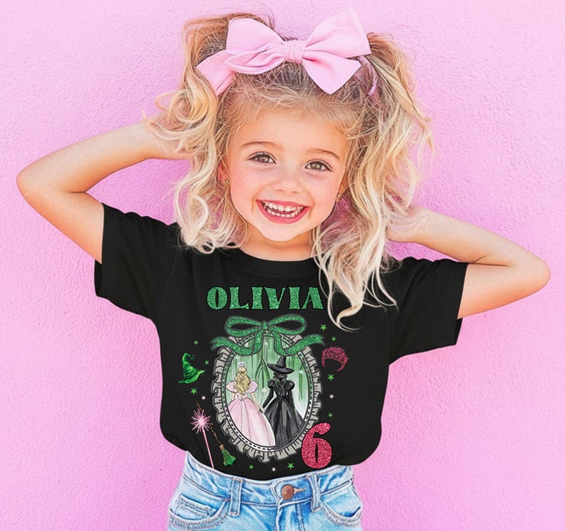 A young girl with blonde hair wearing a black t-shirt with a graphic design and a pink bow, smiling and posing against a pink background.