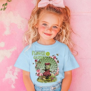 A young girl with blonde hair wearing a blue t-shirt with a cartoon character and floral design, smiling at the camera.