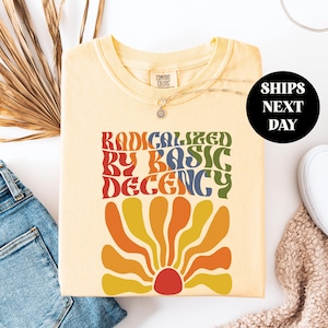May include: A light yellow Comfort Colors sweatshirt with the text "RADICALIZED BY BASIC DECENCY" in a retro font. The design includes a colorful flower graphic in shades of orange, yellow, and red. The sweatshirt is paired with blue jeans and white sneakers.