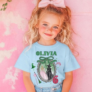 A young girl with blonde hair wearing a blue t-shirt with a graphic design and a pink bow in her hair.