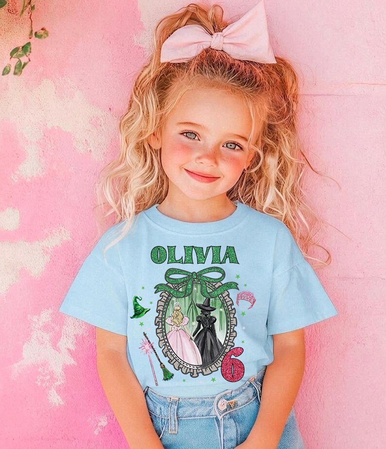 A young girl with blonde hair wearing a blue t-shirt with a graphic design and a pink bow in her hair.
