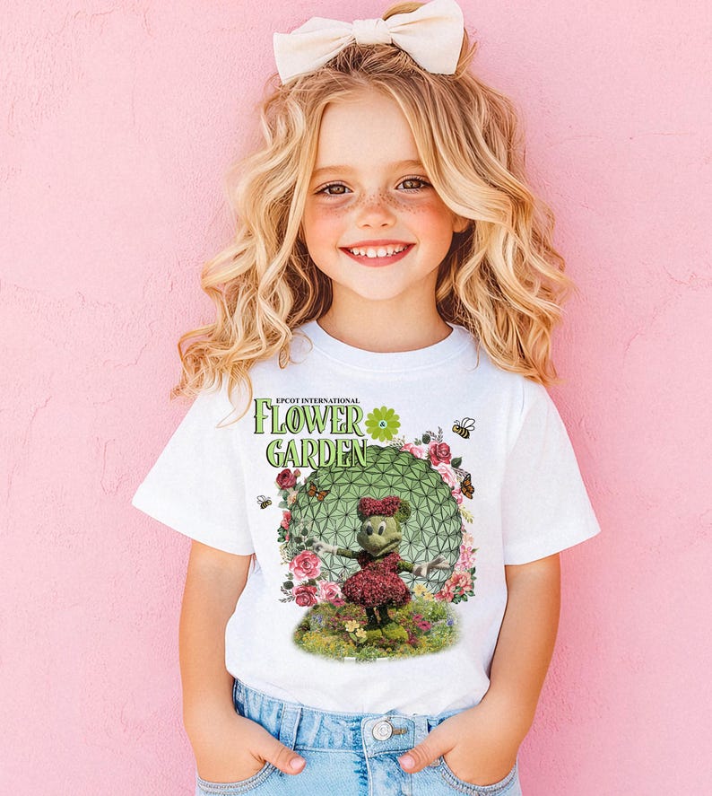 A young girl with curly blonde hair wearing a white t-shirt with a floral design and a bow in her hair, smiling at the camera.