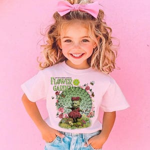 A young girl with curly blonde hair wearing a pink bow and a white t-shirt with a green and pink design of a character from the "Flower Garden" cartoon series.