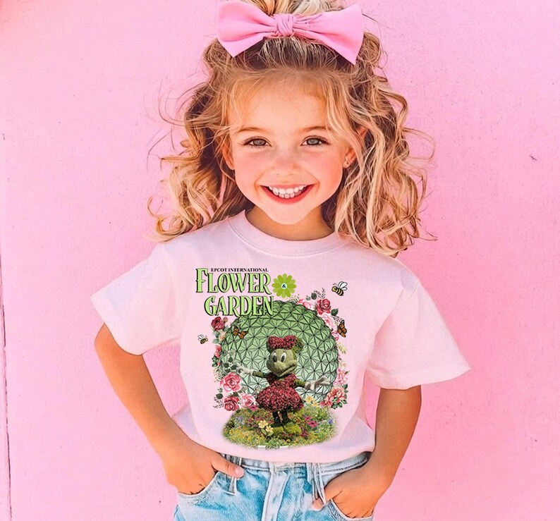 A young girl with curly blonde hair wearing a pink bow and a white t-shirt with a green and pink design of a character from the "Flower Garden" cartoon series.