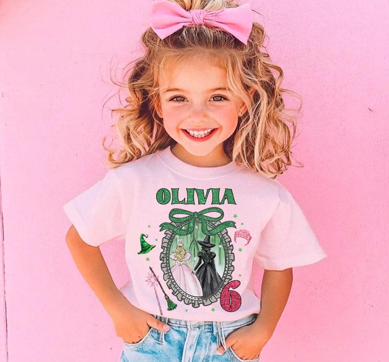 A young girl with curly blonde hair wearing a pink bow and a white t-shirt with a graphic design, smiling at the camera.