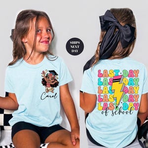 A young girl wearing a light blue t-shirt with a cartoon character and the text "Carol" printed on it. The t-shirt also has a colorful design with the text "LAST DAY OF SCHOOL" printed on the back.